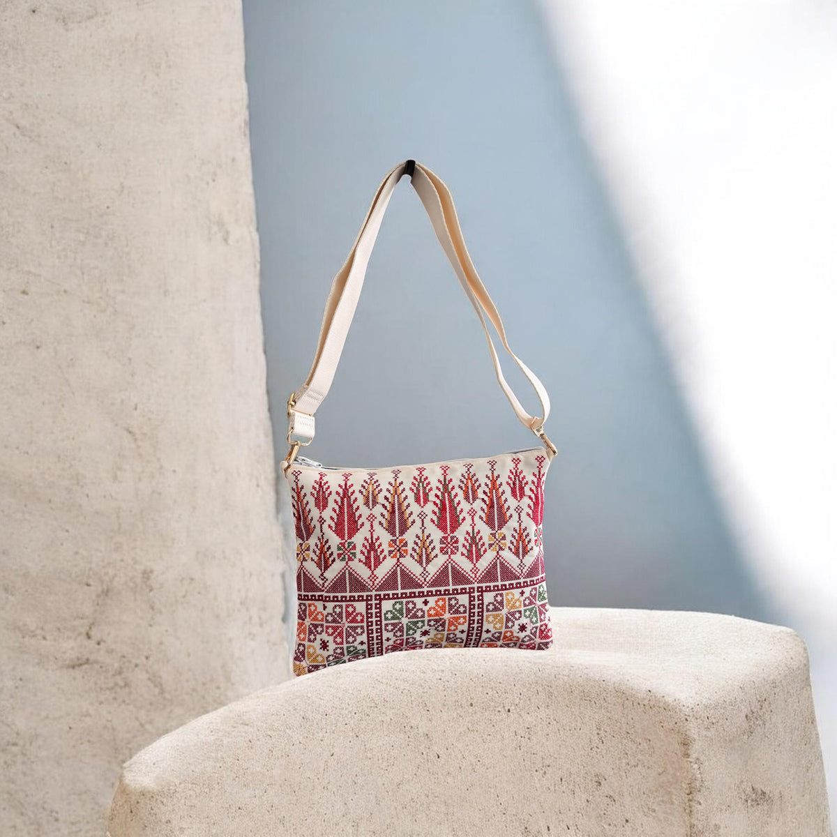 Cypress Tatreez Crossbody hanging by its cream strap, with red cypress motifs and multicolored geometric panels.