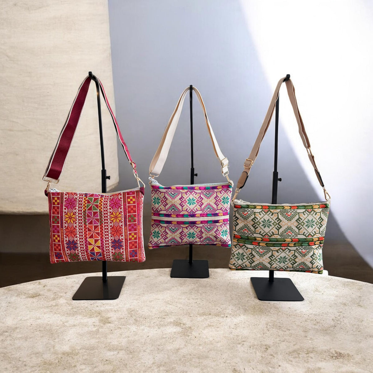 Three Najmeh Crossbody bags in red, purple, and green displayed on stands, showing their embroidered fronts